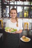 Portrait of smiling young waitress holding fresh Greek salad plate