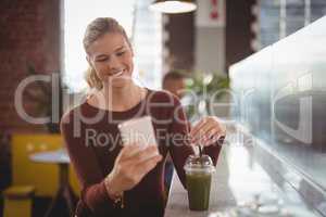Smiling young blond woman using smartphone while sitting with drink at counter