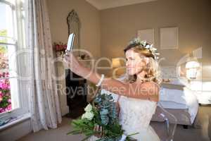 Happy bride taking selfie while sitting on chair in bedroom