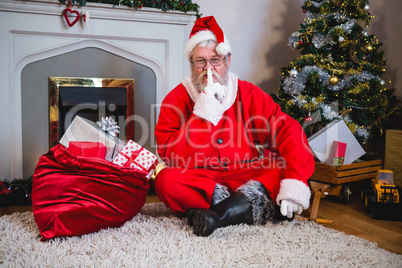 Santa claus with gift sack sitting on rug with finger on lips in living room