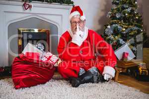 Santa claus with gift sack sitting on rug with finger on lips in living room