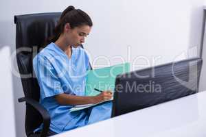Doctor writing on file while sitting on chair against wall
