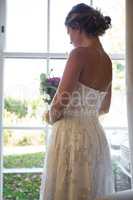 Bride holding bouquet while looking through window at home