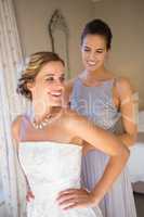 Happy bridesmaid adjusting bride wedding dress in room