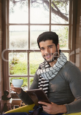 Man wearing scarf with tablet and cup by window in countryside