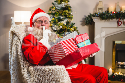 Santa claus with gift boxs sitting on chair with finger on lips in living room