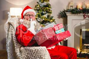 Santa claus with gift boxs sitting on chair with finger on lips in living room