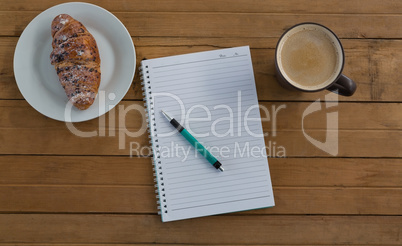 Croissant, coffee, pen, notepad on wooden plank
