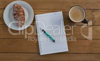Croissant, coffee, pen, notepad on wooden plank