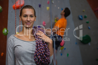 Portrait of athlete carrying rope while standing against man climbing wall in club