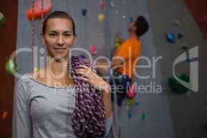 Portrait of athlete carrying rope while standing against man climbing wall in club