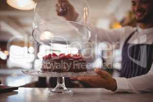 Close up of young waiter holding glass lid over cake on cakestand at counter