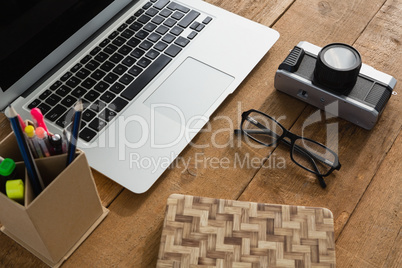 Laptop, camera, spectacles, notepad and pencil holder on wooden plank