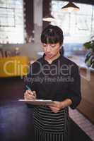 Young attractive female chef writing on clipboard while standing at cafeteria