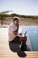 Man using laptop while talking on mobile phone near poolside