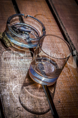 Empty whiskey glass and ashtray with cigarette sticks on wooden table.
