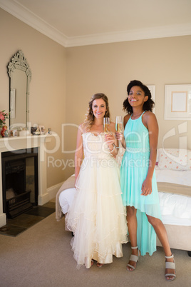 Portrait of happy bride and bridesmaid having champagne while standing in bedroom