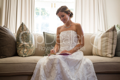 Beautiful bride writing in diary while sitting on sofa at home