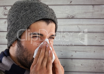 Man against wood with warm hat blowing nose in tissue