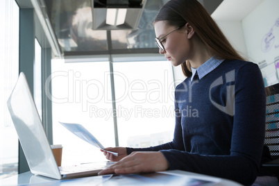 Female executive looking at document wile using laptop