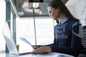 Female executive looking at document wile using laptop