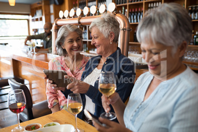 Senior female friends using digital tablet and mobile phone while having wine