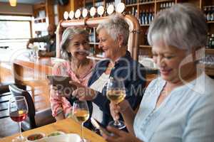 Senior female friends using digital tablet and mobile phone while having wine