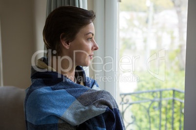 Thoughtful woman wrapped in blanket looking through window