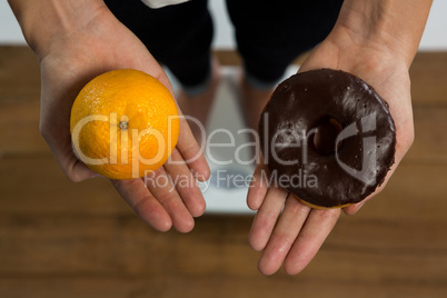 Woman standing on weight machine and holding doughnut and orange