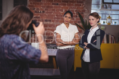 Male photographer clicking picture of confident female colleagues standing with arms crossed