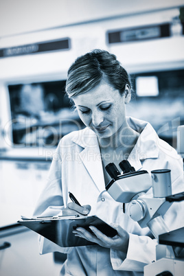 Serious female scientist writing on her clipboard in front of a microscope