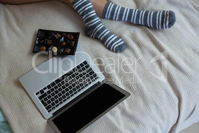 Low section of woman relaxing with chocolates and laptop on bed