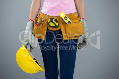 Woman with tool belt and holding hard hat against grey background