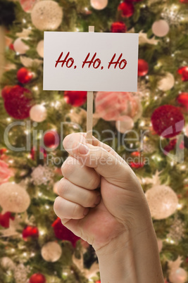 Hand Holding Ho Ho Ho Card In Front of Decorated Christmas Tree.