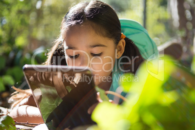Little girl with backpack lying on ground using her tablet on a sunny day in the forest