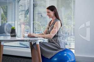 Female executive sitting on exercise ball while using laptop at desk