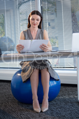 Female executive sitting on exercise ball while reading documents at desk