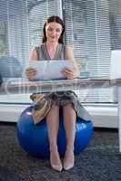 Female executive sitting on exercise ball while reading documents at desk