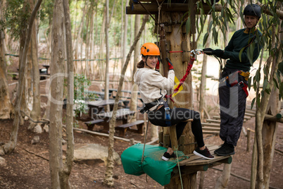 Happy woman leaning on zip line while man standing on wooden platform holding rope