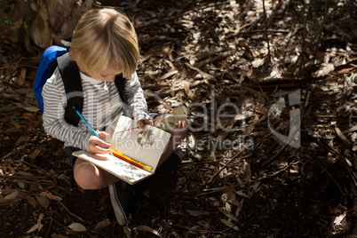 Little girl with backpack sitting on ground writing in notebook