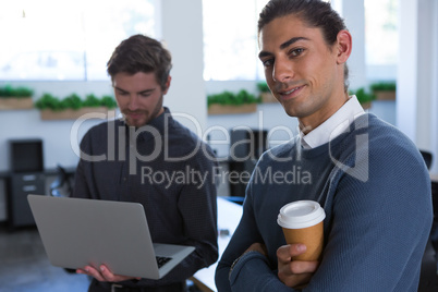 Male executive standing with arms crossed while his coworker using laptop