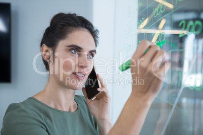 Female executive talking on mobile phone while writing on glass