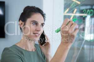 Female executive talking on mobile phone while writing on glass