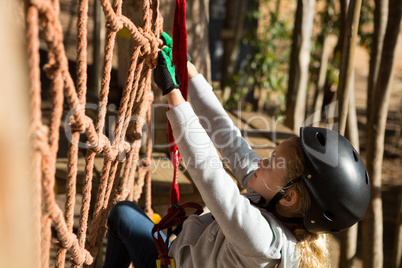 Little girl wearing helmet climbing on rope fence in the forest