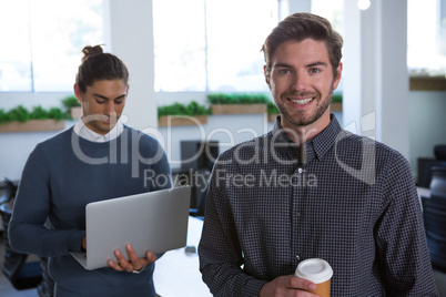 Male executive standing with arms crossed while his coworker using laptop