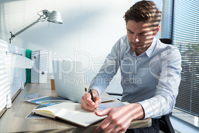 Male executive writing in diary while using laptop