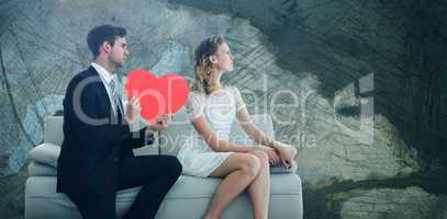 Composite image of man showing red heart shaped paper to woman while sitting on sofa