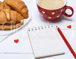empty paper notebook, red cup with coffee and croissants with ch