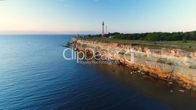 Aerial flying of the drone along the cliffs on the seaside overlooking the lighthouse in the summer at sunset