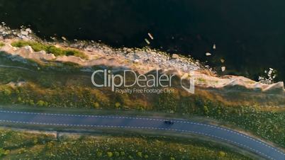 Aerial shot from above driving black car in a field by the rural road near a cliff at the seaside in summer at sunset